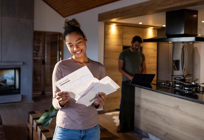A woman standing in a warm, modern kitchen smiles while reading a piece of physical mail, with another person working in the background.
